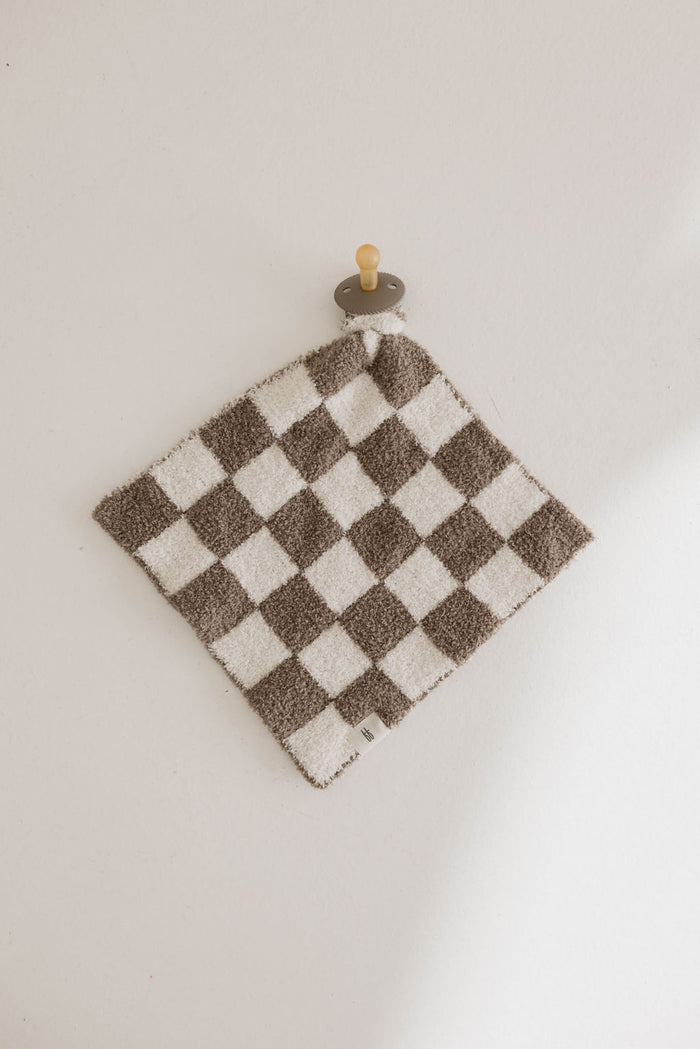 A classic checkerboard print cloth with alternating brown and white squares, named Lovey | Mushroom Checkerboard by forever french baby, hangs on a small, round wooden peg against a white wall. The design appears soft and textured, made from luxurious microfiber feather yarn.