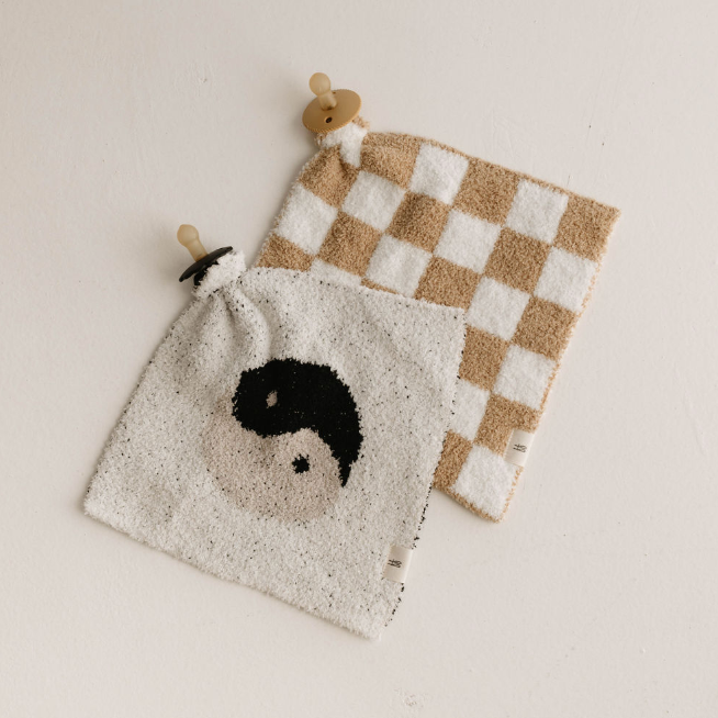 Two soft cloths with pacifiers attached are laid on a light-colored surface. One cloth, named the Lovey | Original Checkerboard from forever french baby, boasts a beige and white checkered pattern made from microfiber feather yarn. The other features an elegant yin-yang symbol in black and beige—perfect little loveys from Forever French Baby.