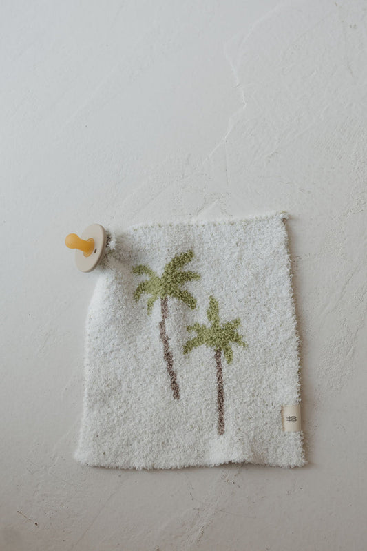 The Lovey | Palm by forever french baby features a yellow pacifier attached to a soft white cloth adorned with two green palm trees, shown lying on a textured white surface.