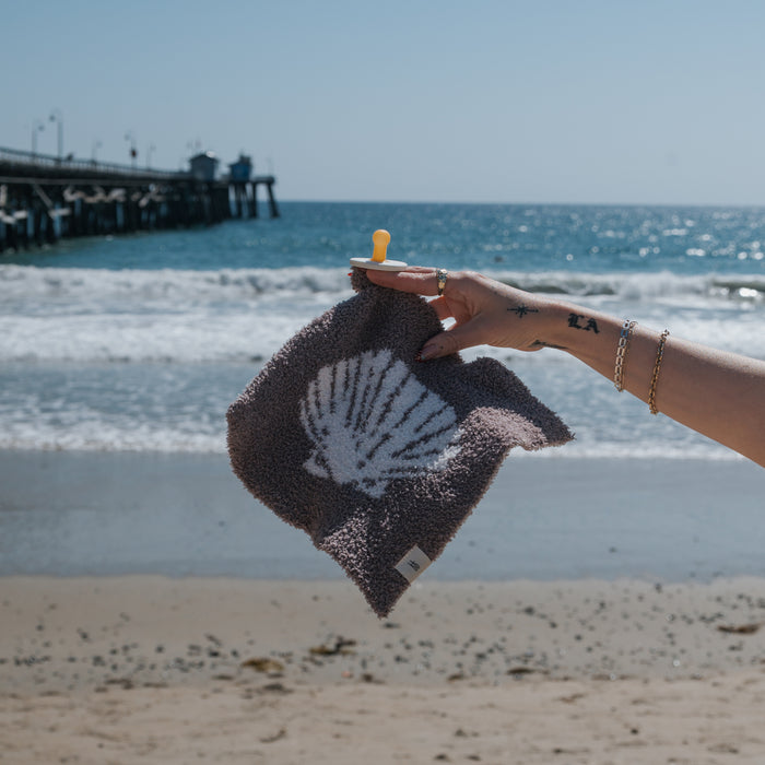 A hand holds the forever french baby Lovey | Seashells, featuring a white seashell print, with rings and bracelets gleaming on a sandy beach as waves roll in near a pier under a clear sky.