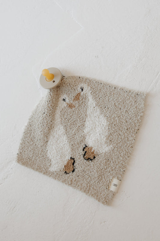 The Lovey | Silly Goose by forever french baby is a soft, square microfiber feather yarn blanket with a cute penguin design, shown on a white textured surface next to a yellow pacifier for an extra cozy touch.
