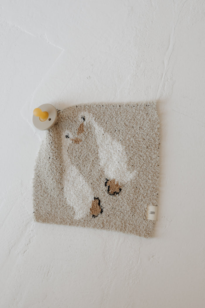 The Lovey | Silly Goose by forever french baby, a beige microfiber feather yarn towel with a white duck design, hangs on a yellow hook against a white wall.
