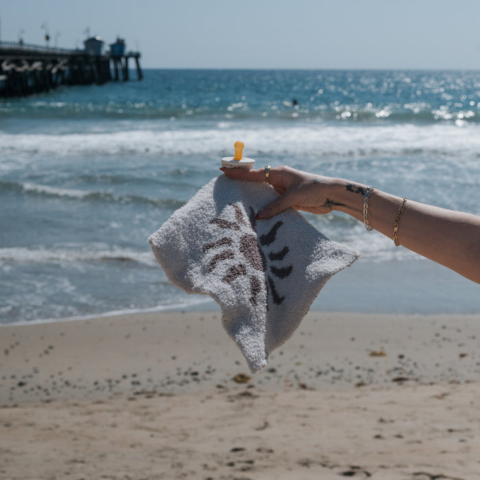 A hand holds the forever french baby Lovey | Summer Sun—a soft microfiber blanket with a cheerful sun print and attached yellow pacifier—against an ocean backdrop with sandy beach and pier.