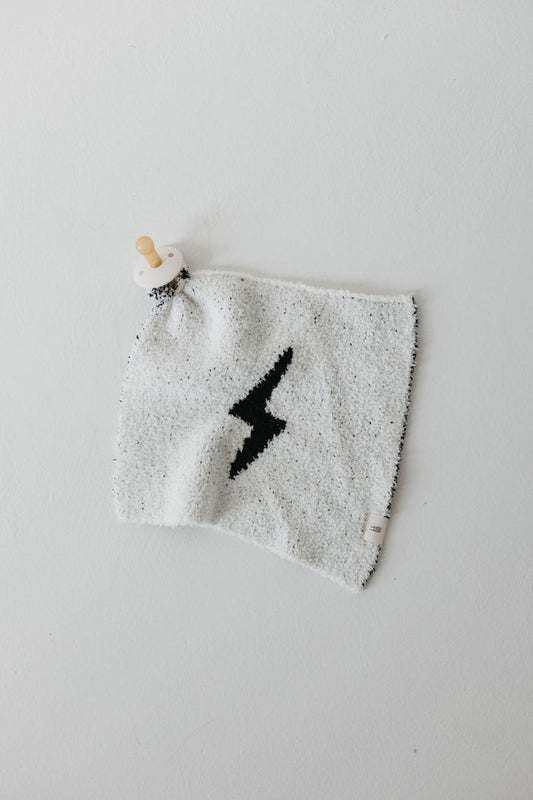 The "Lovey | Black & White Lightning Bolt" by Forever French Baby is a soft, white square blanket made from microfiber feather yarn. It features a black lightning bolt design in the center, with a wooden knob attached to one corner and a small tag sewn into one edge. This makes it perfect as a lovey for your little one.