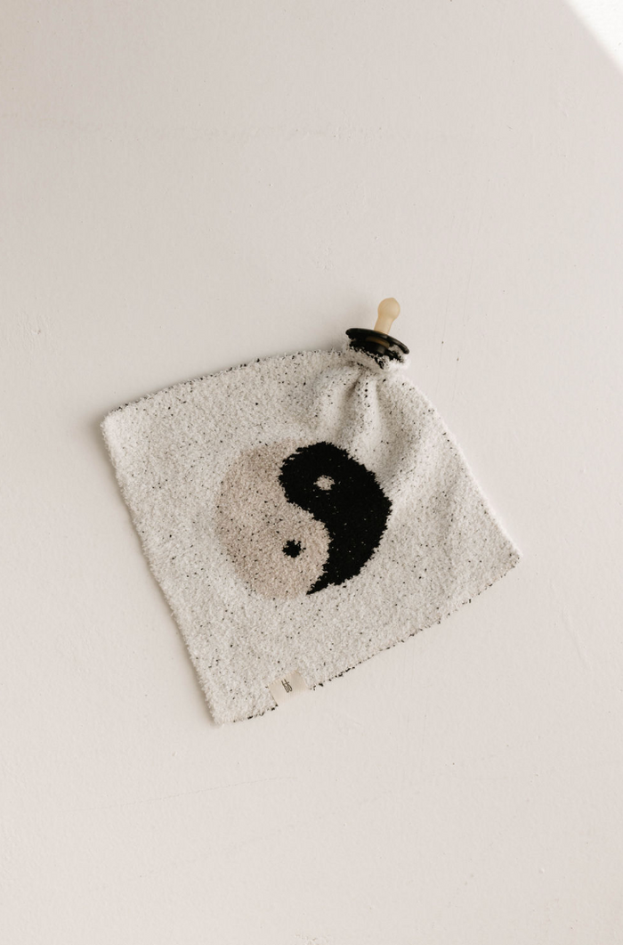 The Lovey | Yin-Yang Black & White from forever french baby is a square-shaped knitted cloth made from microfiber feather yarn, featuring a black and white yin-yang symbol at its center and a pale-colored pacifier attached to one corner. This softest lovey rests on a plain, light-colored surface.