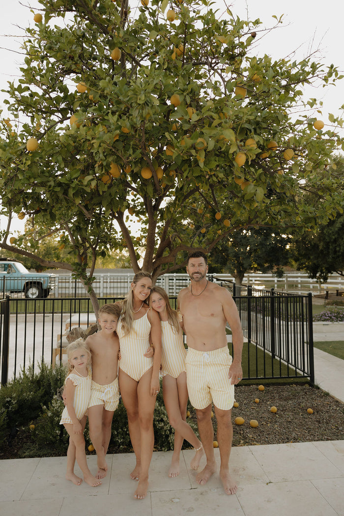 A family of five in matching yellow-and-white striped swimsuits, including the limited edition Sun Rays Child Board Short by lolo webb 2, smiles together outdoors in front of a lemon tree near a fenced yard.