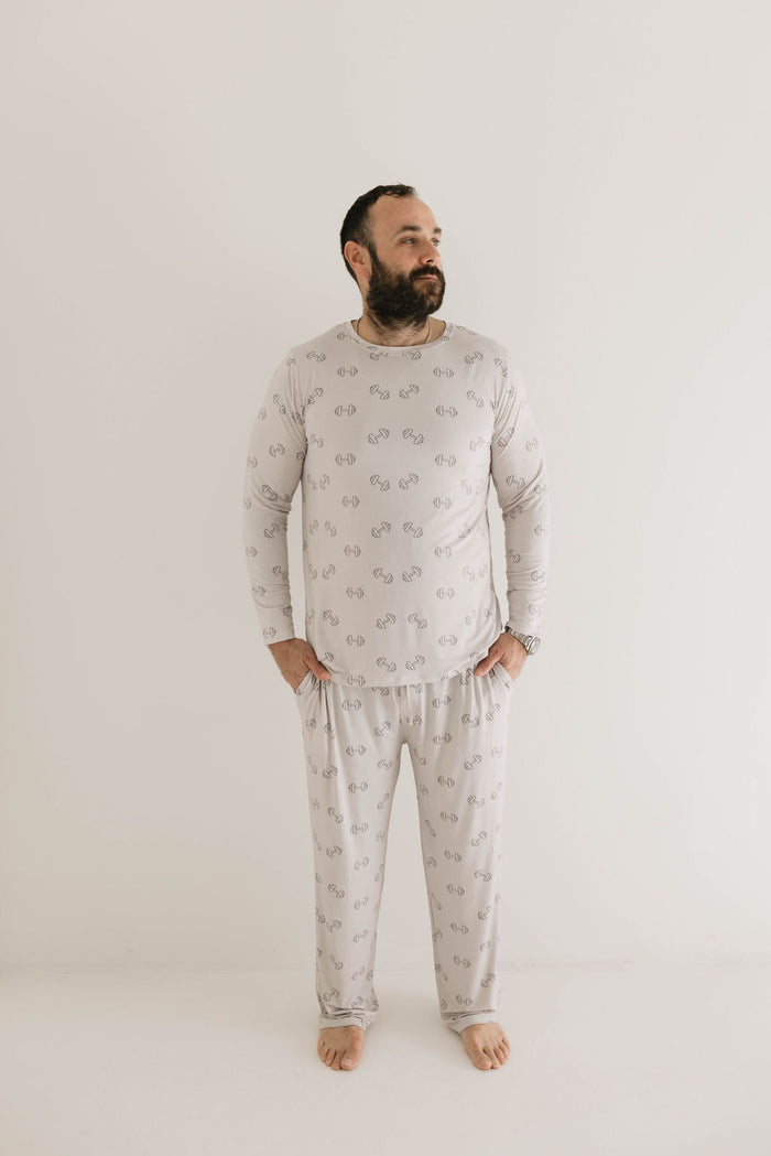 A bearded man stands barefoot in a neutral-colored room, wearing matching Adult Long Sleeve Bamboo Pajamas | Arm Day by forever french baby, adorned with a delicate pattern of small dumbbell weights. He gazes serenely to the side, looking ready for a comfortable night's sleep, with his hands resting on his hips.