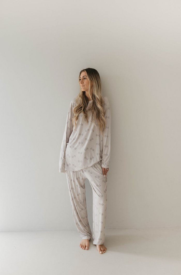 A woman with long, wavy hair stands against a plain white wall. She is wearing light-colored, loose-fitting "Adult Long Sleeve Bamboo Pajamas | Arm Day" by forever french baby, which feature a subtle pattern. She is barefoot and looking off to the side with a relaxed expression, clearly ready for a comfortable night's sleep.