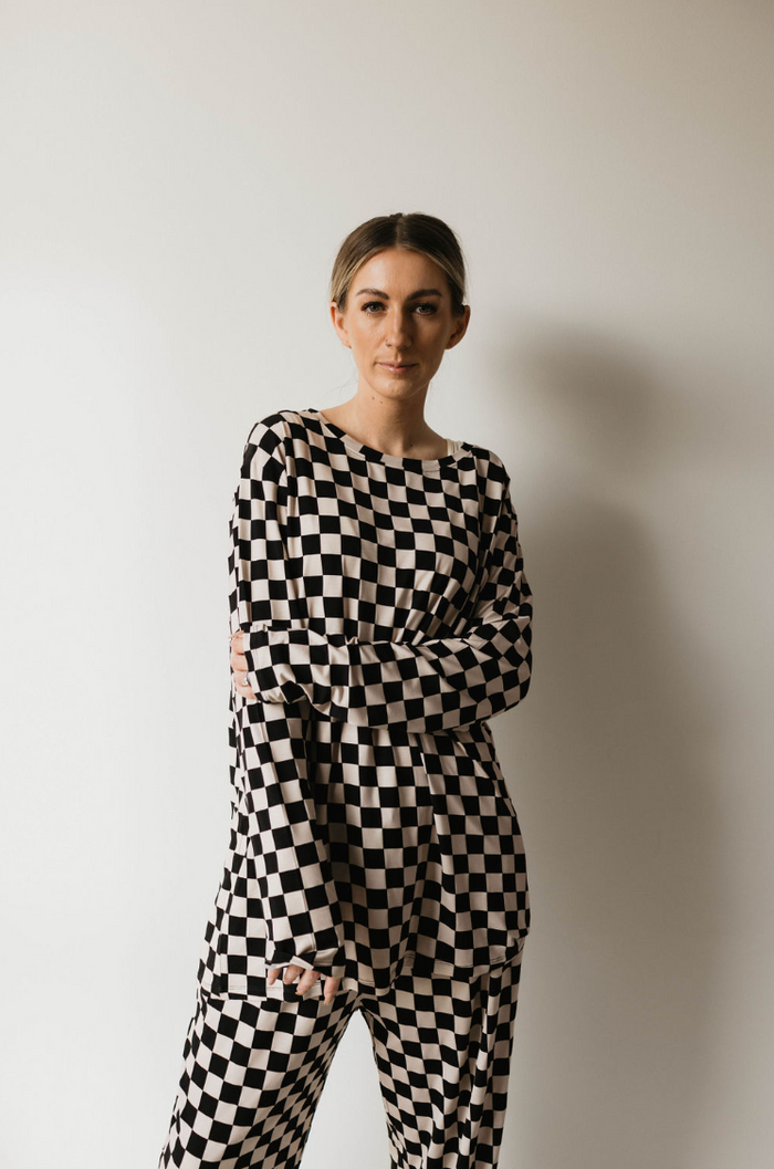 A woman stands against a plain white background, wearing the Adult Bamboo Pajamas in a black and white checkerboard pattern by forever french baby. She has her hair pulled back and is staring directly at the camera with a neutral expression, one arm crossed over her body.