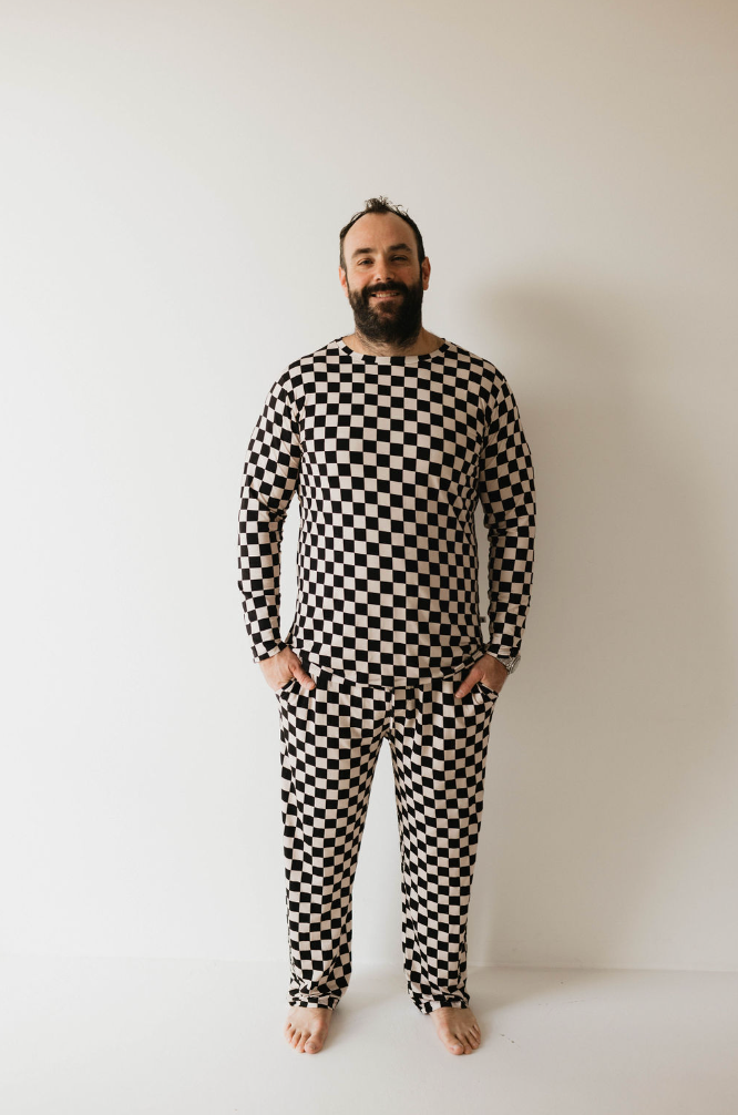 A bearded man stands against a plain white background, wearing forever french baby's Adult Bamboo Pajamas in a black and white checkerboard pattern. He is barefoot, has his hands relaxed by his sides, and is smiling at the camera.