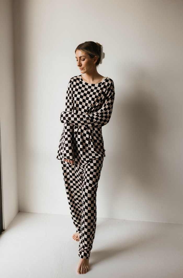 A person stands barefoot in a minimalistic, light-filled room, wearing an Adult Bamboo Pajamas set in Black & White Checkerboard by forever french baby. With a relaxed expression, their arms are loosely crossed as they lean slightly against the wall behind them.