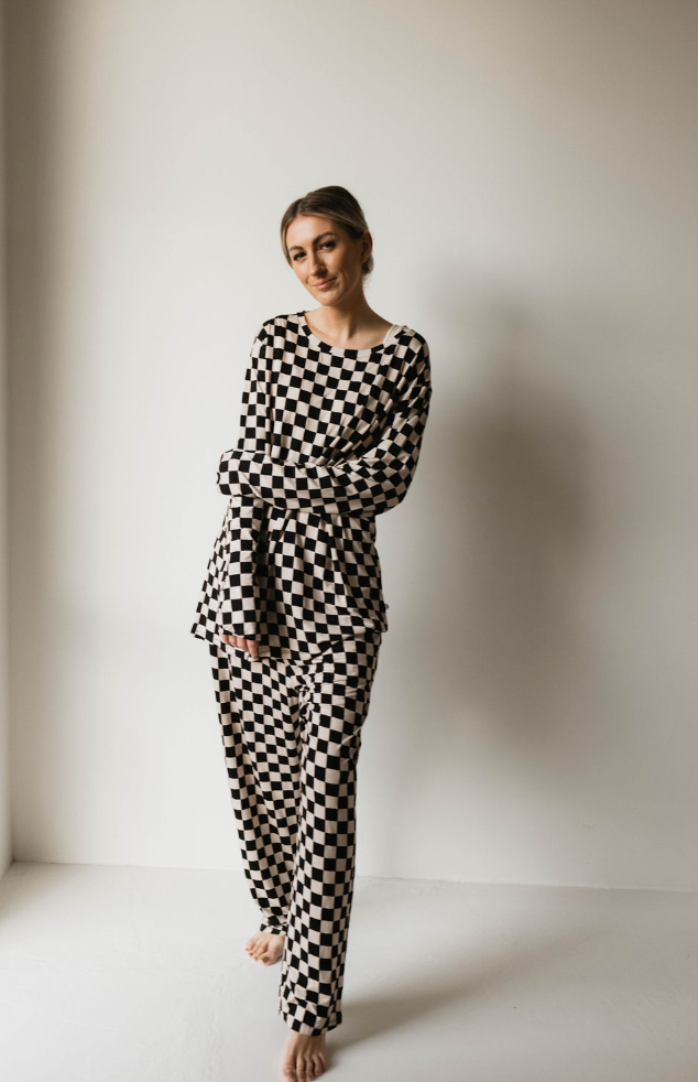 A woman stands barefoot against a plain white wall, wearing Adult Bamboo Pajamas in a black and white checkerboard pattern. She is smiling gently and tilting her head slightly to one side, with her arms loosely crossed over her torso, looking as stylish as pieces from the forever french baby collection.
