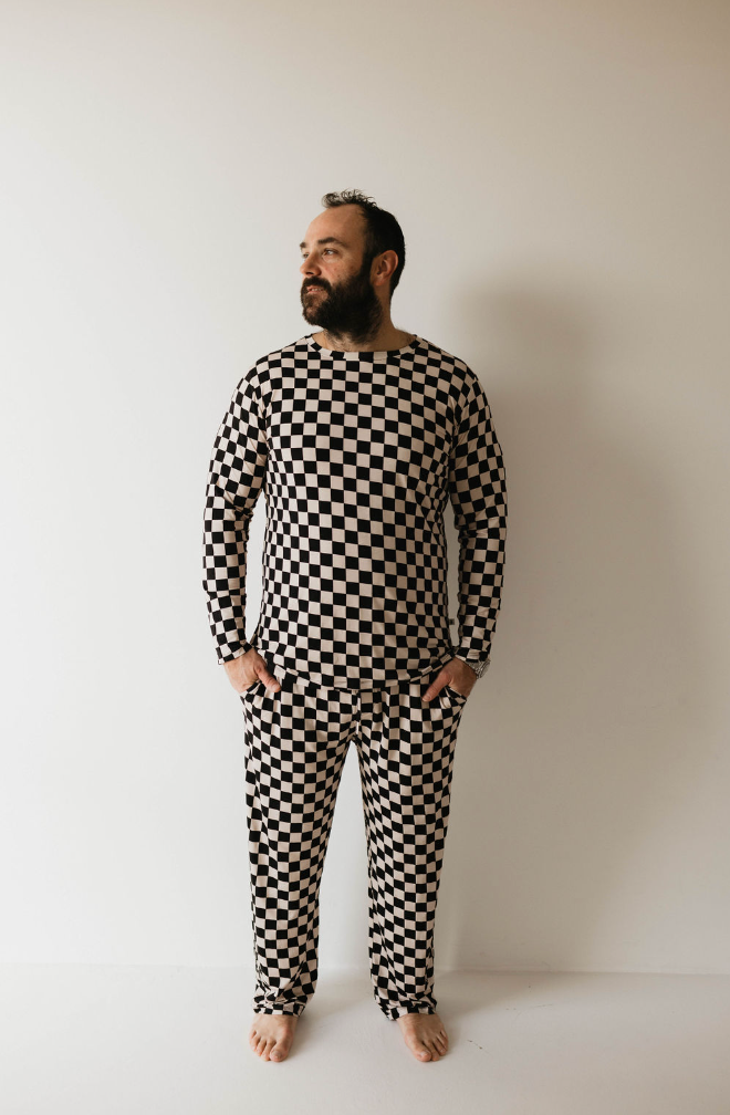 A man with a beard stands barefoot against a white wall, wearing forever french baby's Adult Bamboo Pajamas in Black & White Checkerboard. His hands are in his pockets, and he looks to the side.