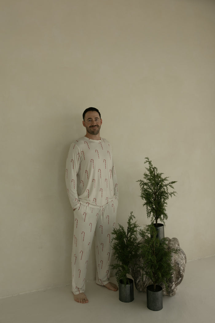 A man smiles barefoot beside green potted plants, wearing forever french baby's Men's Bamboo Pajamas | Candy Cane—a cream set with red candy canes—standing against a beige wall.