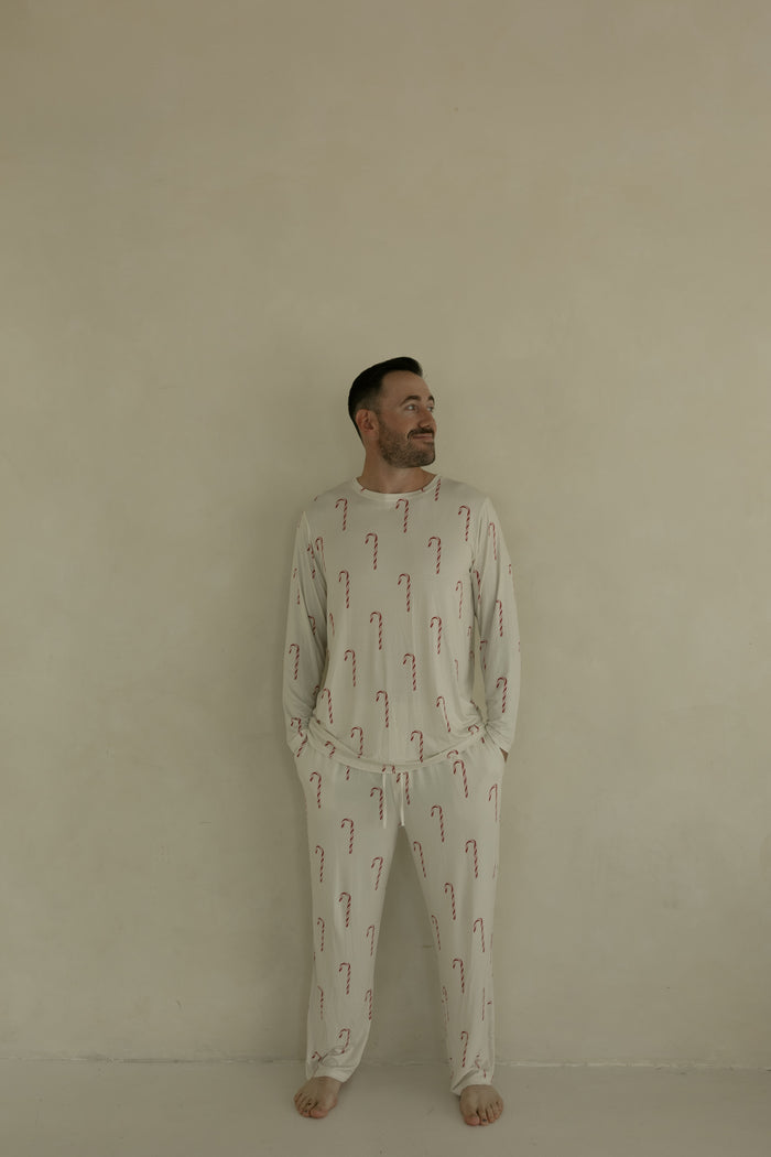 A man stands barefoot against a plain beige wall, wearing forever french baby Men's Bamboo Pajamas | Candy Cane, featuring a light-colored fabric with red candy cane pattern. He has his hands in his pockets and is smiling slightly while looking to the side.