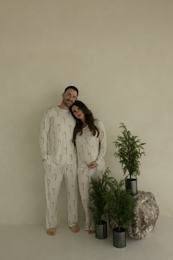 A smiling couple stands barefoot indoors, the man wearing forever french baby's Men's Bamboo Pajamas | Candy Cane. The woman cradles her baby bump beside three small potted evergreens against a plain light wall.