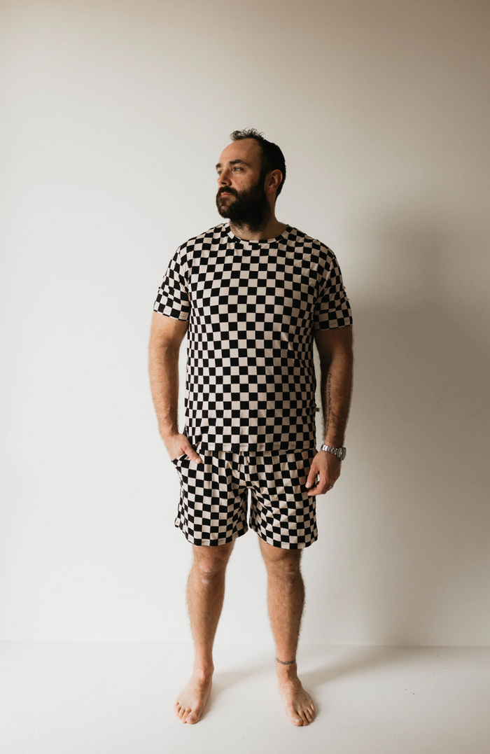 A man stands barefoot against a plain white background, wearing the forever french baby Adult Bamboo Short Pajamas in Black Checkerboard. He is looking off to the side, with his hands relaxed by his sides. He has a beard and a watch on his left wrist.