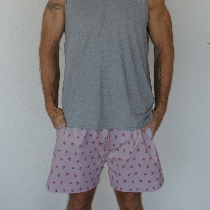 A man models the Men's Board Short | Matilda by forever french baby, featuring a pink strawberry print and gray sleeveless shirt, standing against a plain white wall with his hands in his pockets. His face is not visible.