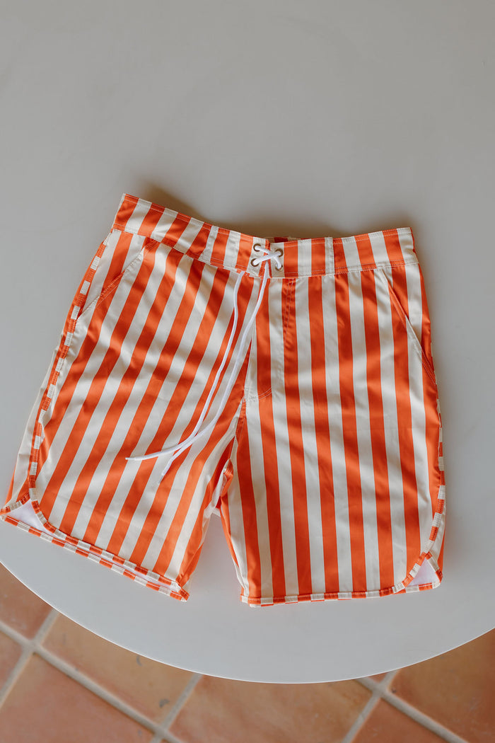 The Men's Boardshort | Poolside by forever french baby, featuring orange and white vertical stripes and a drawstring waist, is laid flat on a round, light-colored table.