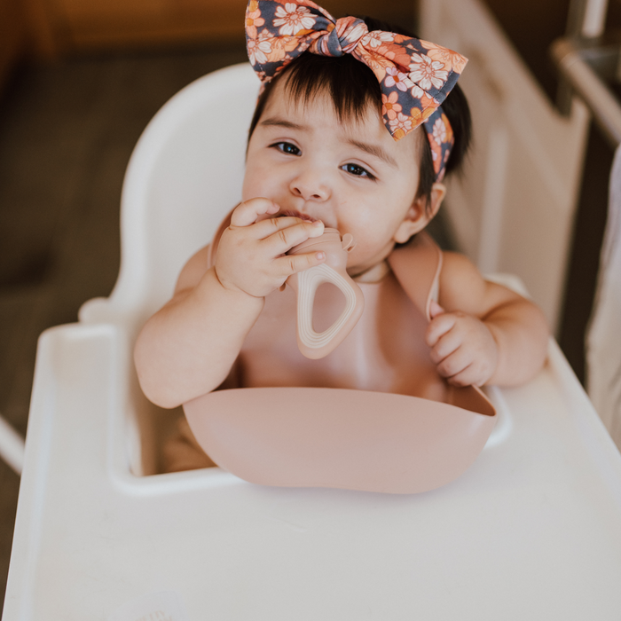 A baby with a floral headband sits in a high chair, holding a Modern Mod Feeder from forever french baby. The baby is wearing a bib and has a playful expression. The setting appears to be a home kitchen.