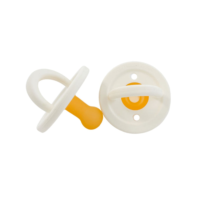 Two Baby Essentials are shown in the image against a white background: Modern Pacifiers in Coconut Milk from forever french baby. One lies flat with its handle facing upward, while the other, featuring a sleek silicone design, is placed upright with the nipple facing downward.