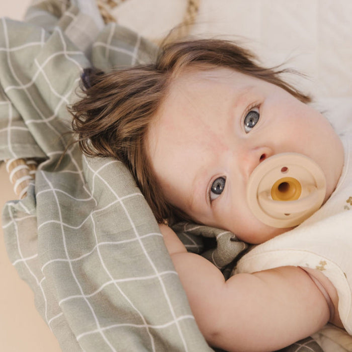 A baby with light skin and brown hair is lying on a plaid blanket. The baby has wide-open blue eyes and is using the Modern Pacifier | Coconut Milk from forever french baby. The scene appears warm and cozy, showcasing essential comfort for your little one.