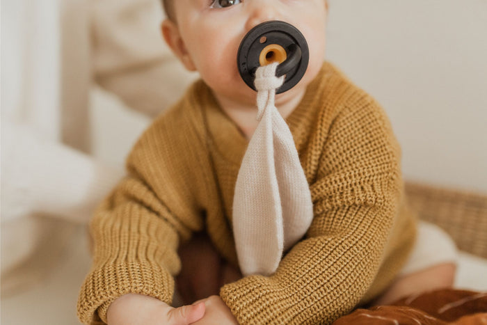 A baby with a pacifier in its mouth sits wearing a knitted brown sweater. The Modern Pacifier in Coconut Milk by forever french baby, designed with silicone for safety, is attached to a cloth. The baby is partially holding the cloth with its hand. The neutral background highlights the baby's attire and baby essentials perfectly.