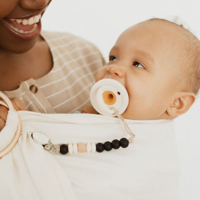A close-up image shows a smiling adult and a baby. The baby, wrapped in soft, white fabric, holds a Modern Pacifier in Coconut Milk from forever french baby. The adult, partly visible in a beige and white striped shirt, securely cradles the baby. Nearby dangles a beaded pacifier clip from the Baby Essentials collection.