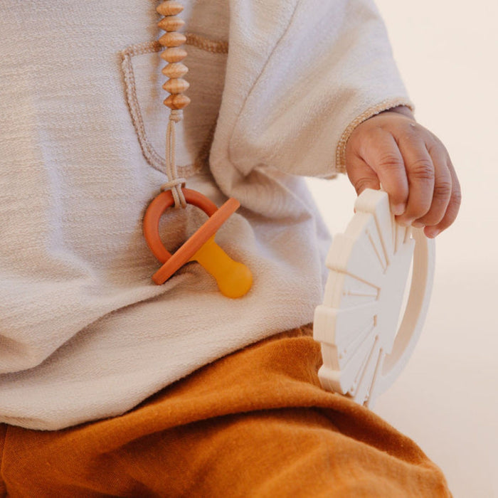 A child wearing a white knitted sweater and brown pants is holding a white toy wheel with one hand. A Modern Pacifier | Coconut Milk from forever french baby, featuring an orange handle, is attached to their sweater with a beaded clip. The background is neutral and out of focus.