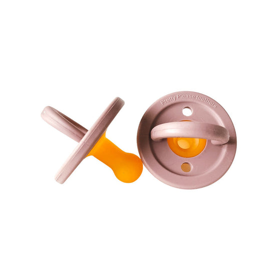Two baby pacifiers are shown. Both feature a soft rubber nipple in fawn color, essential for any Baby Essentials kit. The exterior plastic parts are in a muted pink tone. One Modern Pacifier by forever french baby is lying on its back, revealing the nipple, while the other lies face up, showing the handle.