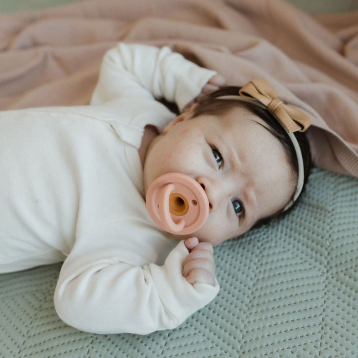 A baby wearing a white onesie lies on a light green textured blanket. The baby has a tan headband with a bow and is sucking on a Modern Pacifier in Mahogany Rose by forever french baby while looking up with a curious expression. A pink blanket is partially visible in the background.
