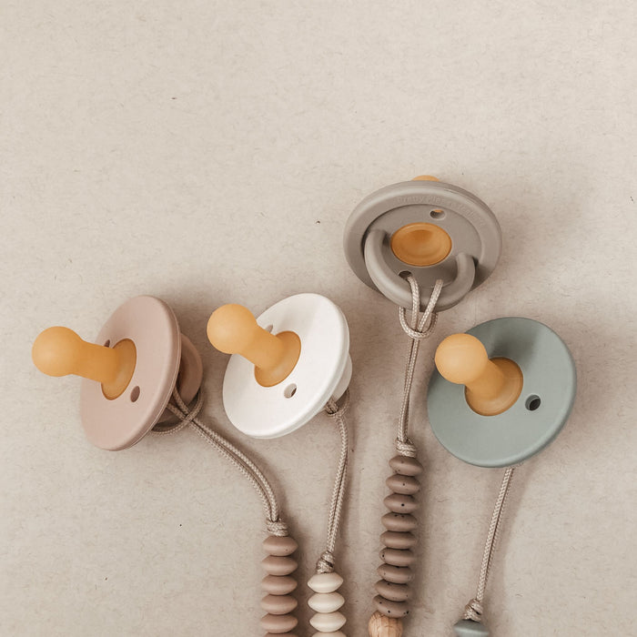 Four "Speckled Almond" Modern Pacifiers by forever french baby, featuring muted pastel colors—beige, white, gray, and light green—are laid out in a row on a neutral background. Each pacifier is attached to a beaded clip in matching tones, making them perfect baby essentials.