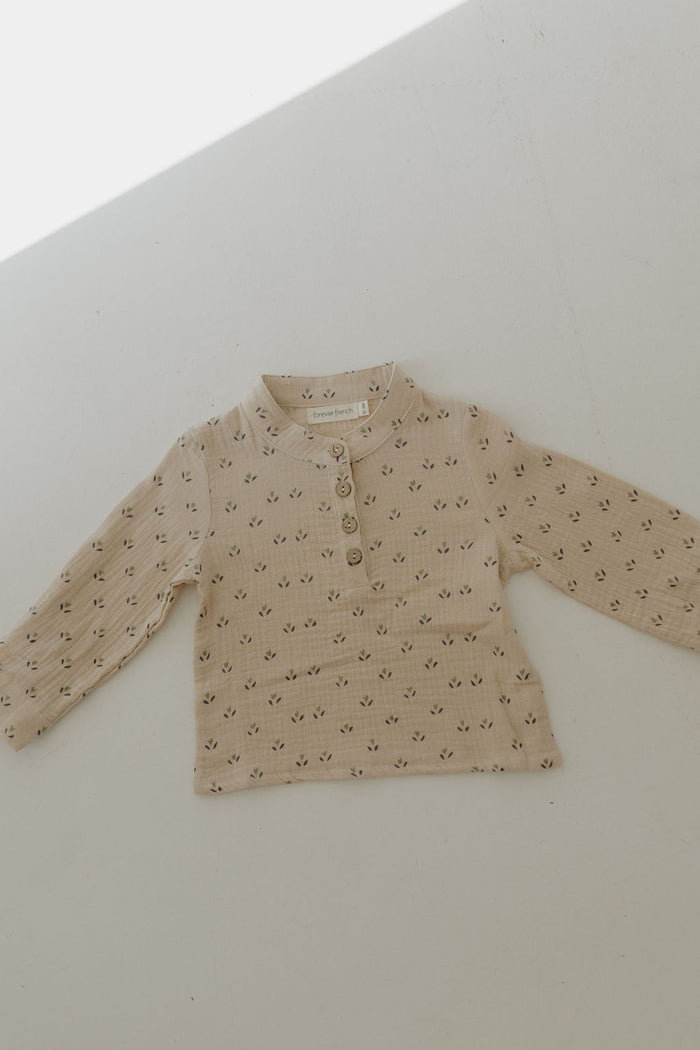 The Muslin Button Up | Flora by forever french baby is a beige, long-sleeved muslin cotton shirt featuring dark floral patterns, a round collar, and three buttons—perfect for adding charm to your kids' clothing collection.