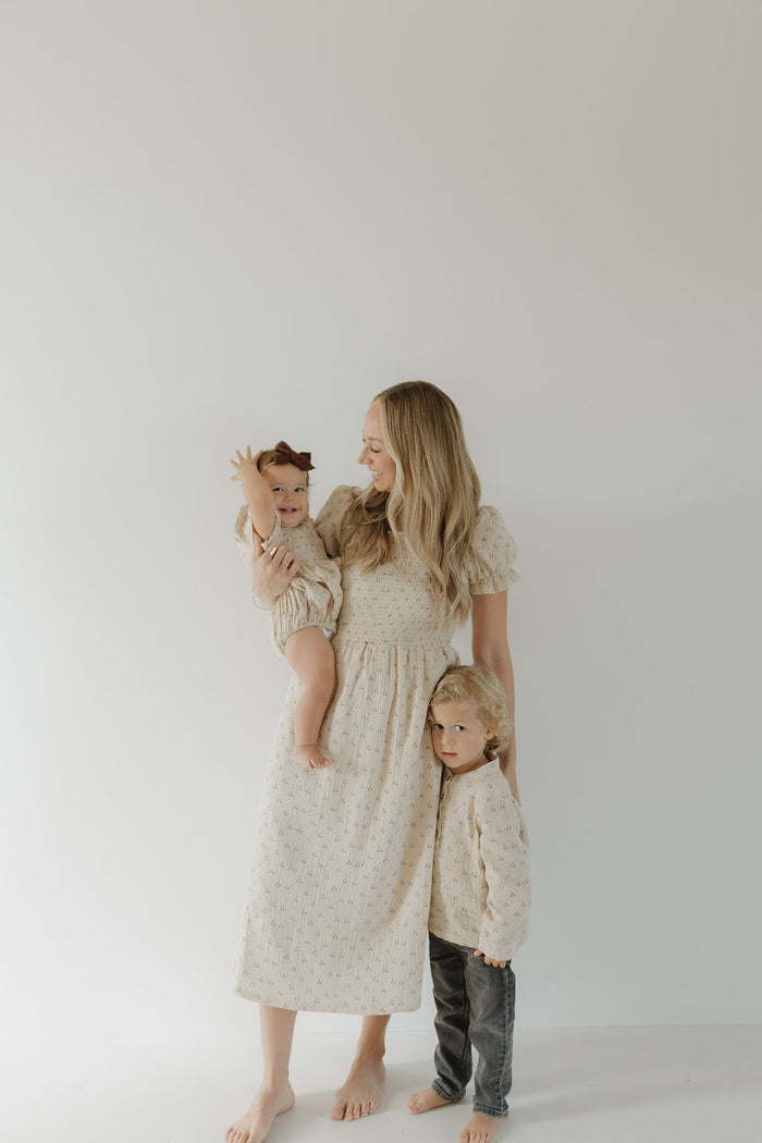 A barefoot woman smiles at her children as they all wear matching forever french baby Flora Women's Short Sleeve Muslin Dresses, posing together against a plain light background in soft, cream-colored muslin fabric.