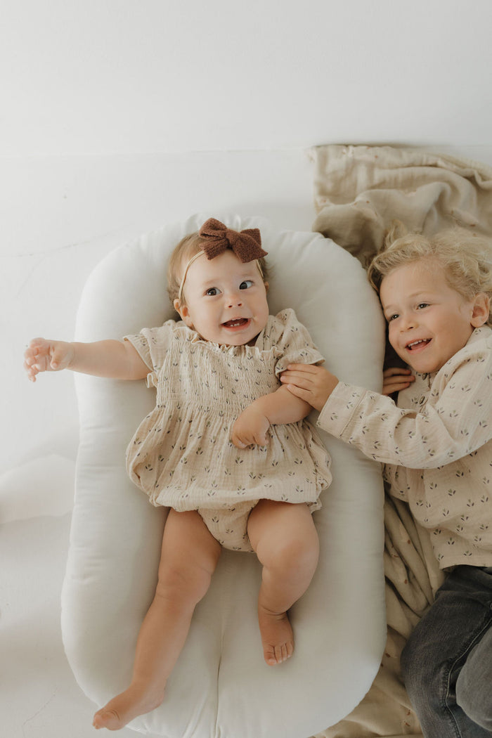 A baby in a beige muslin cotton dress and brown bow lies smiling on a cushion. Next to her, a young child wears the Muslin Button Up | Flora by forever french baby and smiles while holding the baby's hand on a soft blanket.