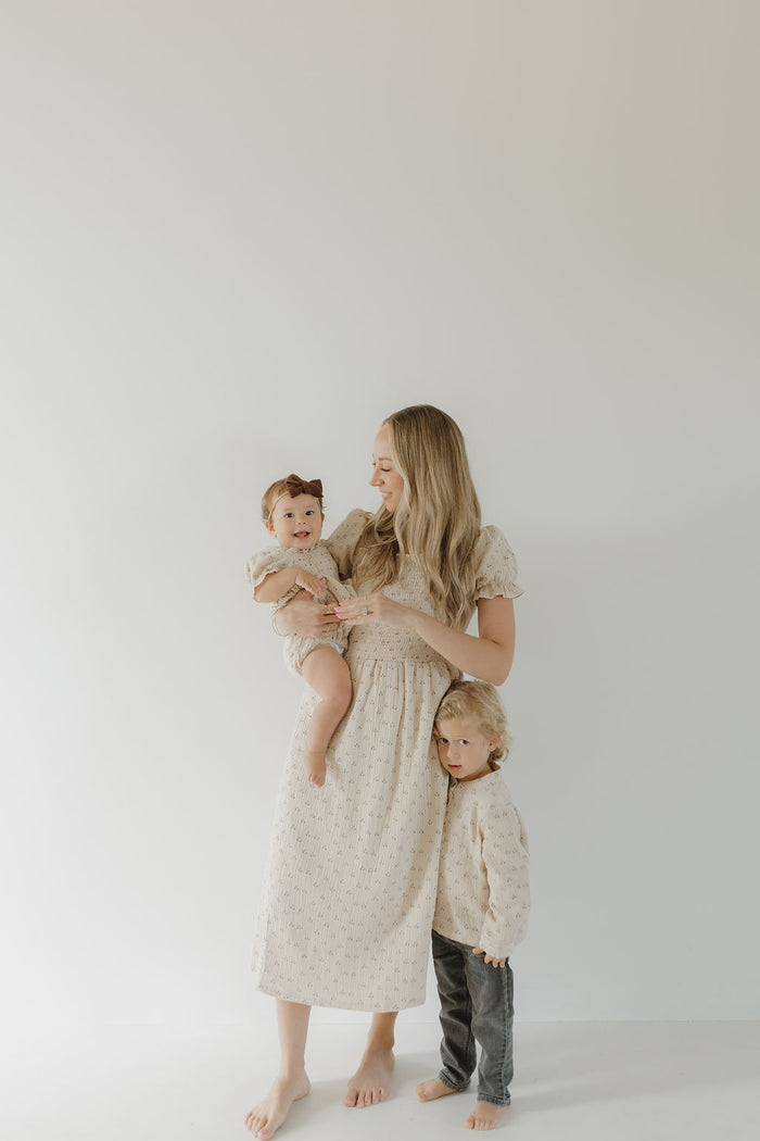 A woman wears the forever french baby Women's Short Sleeve Muslin Dress | Flora, standing barefoot against a white wall, holding a baby with a brown bow beside a small child in a cream sweater and gray pants. All face forward with soft expressions.