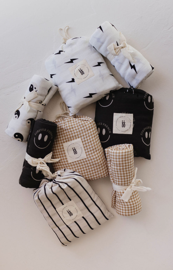 A collection of neatly folded baby blankets and swaddles in various patterns, including gingham, stripes, lightning bolts, yin-yang symbols, and smiley faces. Each is tied with ribbon and arranged on a light surface alongside the elegant Muslin Crib Sheet | Black & Ivory Yin Yang by forever french baby. Tags with the "bb" logo are visible.