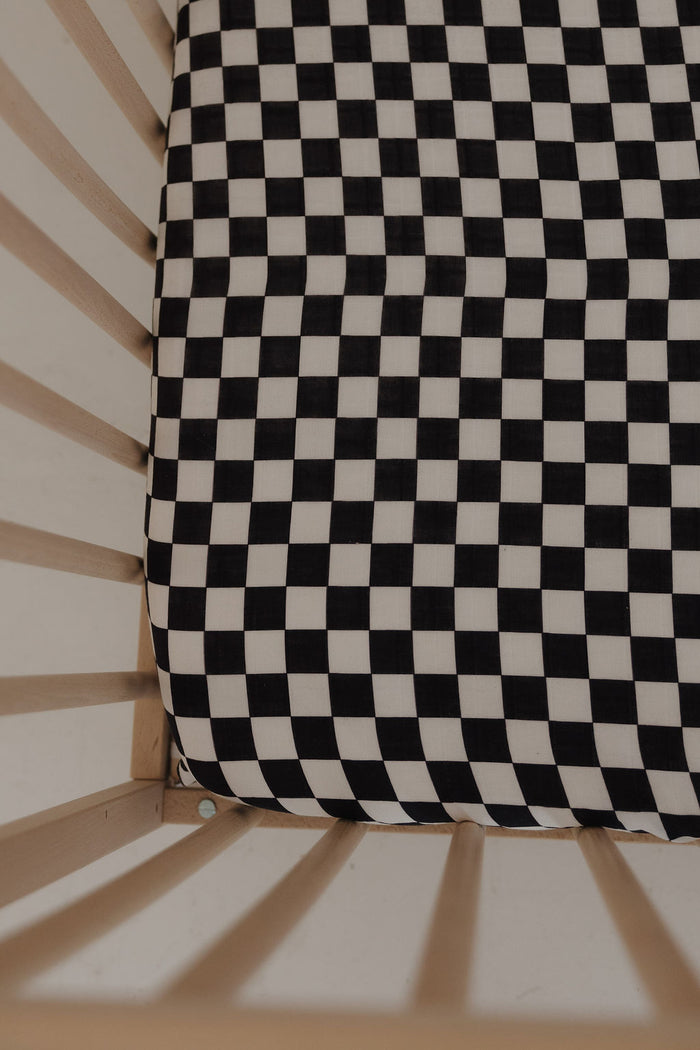 A close-up of a crib features the forever french baby Muslin Crib Sheet | Black & White Checkerboard, crafted from soft cotton muslin and framed by light wooden slats—an elegant addition to your baby's bedding.