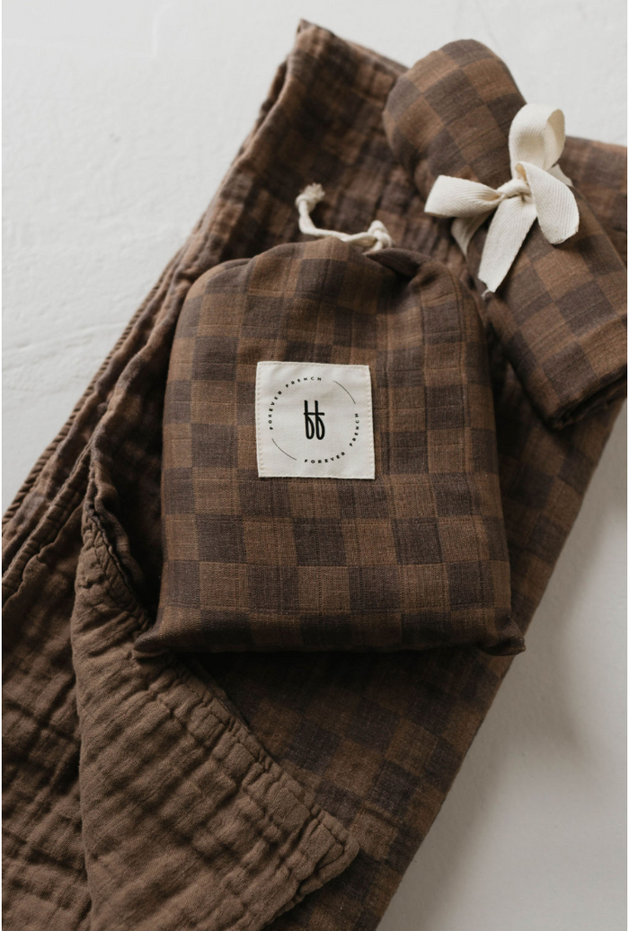 A set of folded brown plaid textiles, including a quilted piece and a small drawstring bag, all crafted from soft muslin cotton and featuring a stitched tan label with the initials "bb" in lowercase letters enclosed in a dotted circle. Among them is the "Muslin Crib Sheet | Faded Brown Checkerboard" from forever french baby, neatly arranged on a white surface.