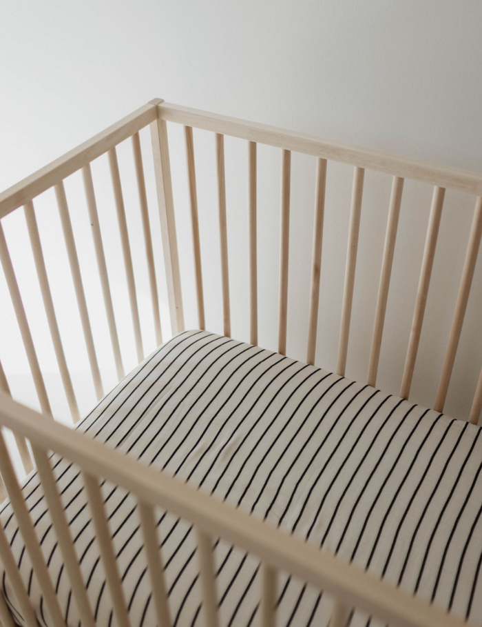 A wooden baby crib with vertical slats holds the Muslin Crib Sheet | Grey & Black Stripe from forever french baby. The crib is set against a plain white wall in a brightly lit room.