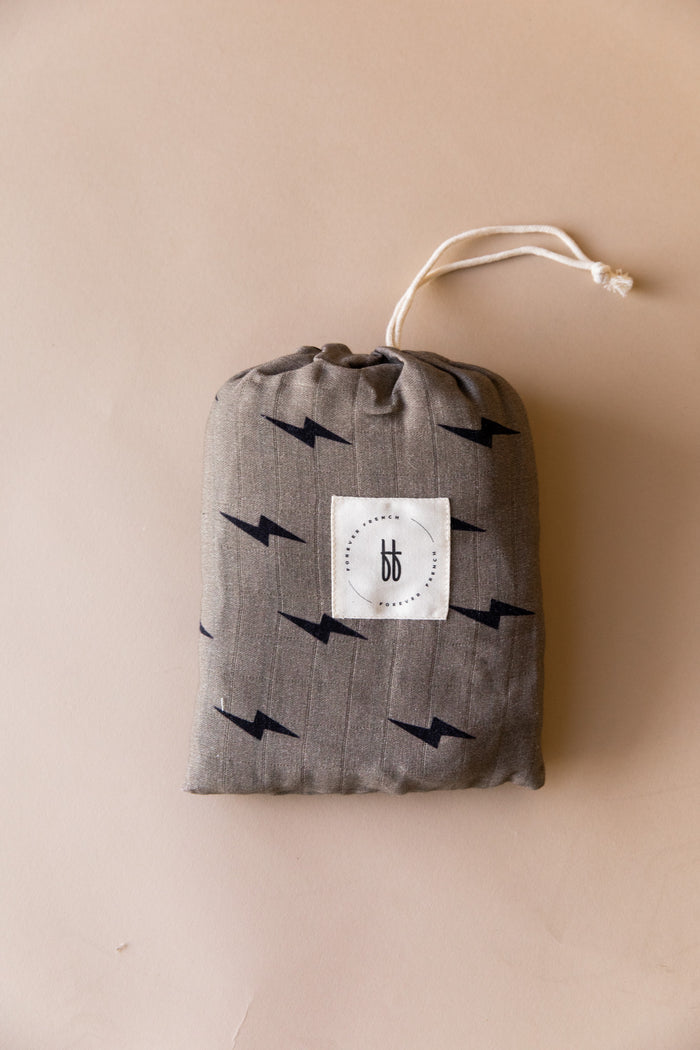 Crafted from muslin cotton, the "Muslin Crib Sheet | Lightning Bolt" by forever french baby showcases a striking black lightning bolt pattern. This beige crib sheet features a centrally placed white square label with black text and the brand's logo, seamlessly blending style with simplicity against its plain background.