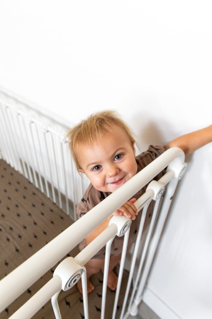 A smiling toddler with short hair holds onto the rail of a white metal crib. Standing inside on a patterned mattress adorned with the Muslin Crib Sheet | Lightning Bolt by forever french baby, the child looks up at the camera. The background features a plain white wall.