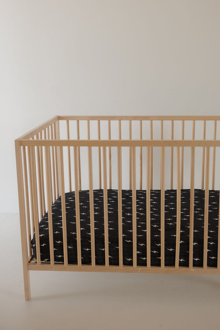 A minimalist wooden crib with a black mattress, topped with the forever french baby Muslin Crib Sheet | Midnight Bolt, sits against a plain light wall in an uncluttered room.