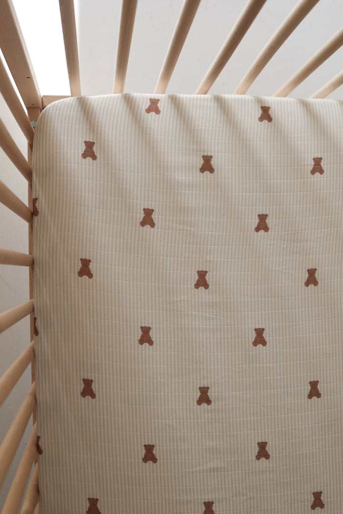 The forever french baby Muslin Crib Sheet | Teddy features a cute brown teddy bear design on subtle stripes, shown here on a light wood crib. Soft light enhances the cozy atmosphere—perfect for your baby's bedding.