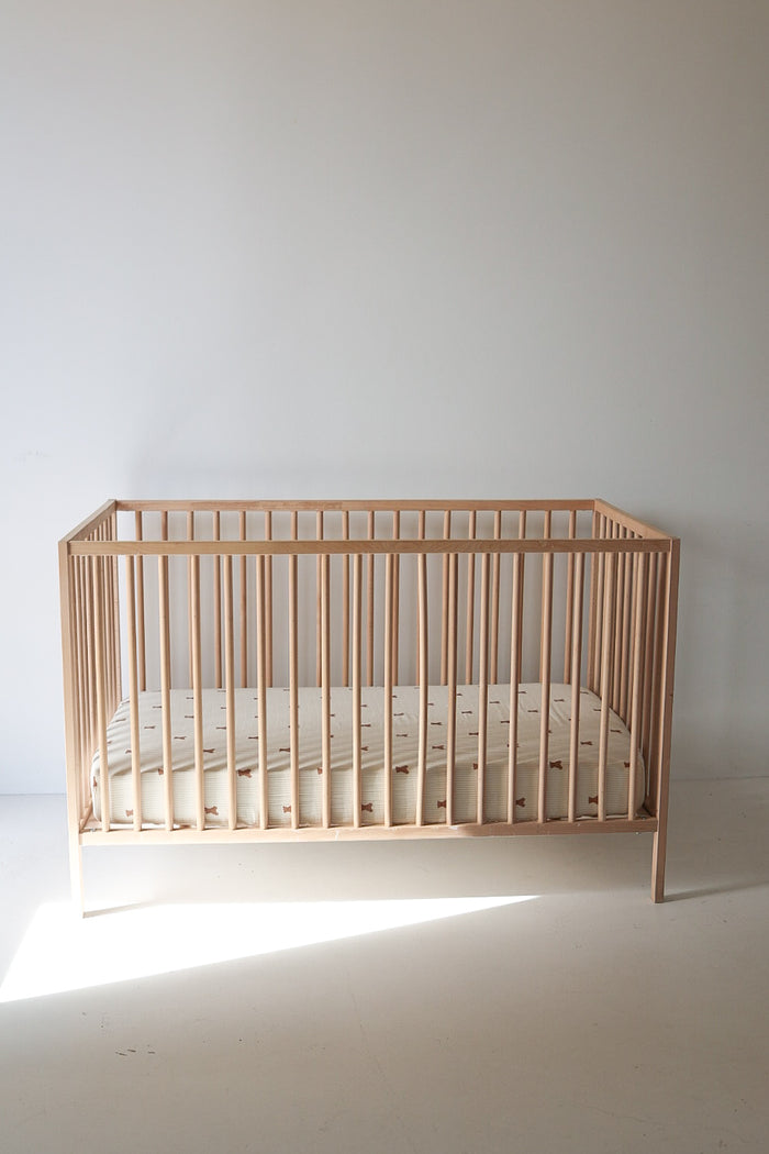 A minimalist wooden crib with vertical slats features the forever french baby Muslin Crib Sheet | Teddy, its subtle light pattern standing out against a plain white wall in a bright, empty room.