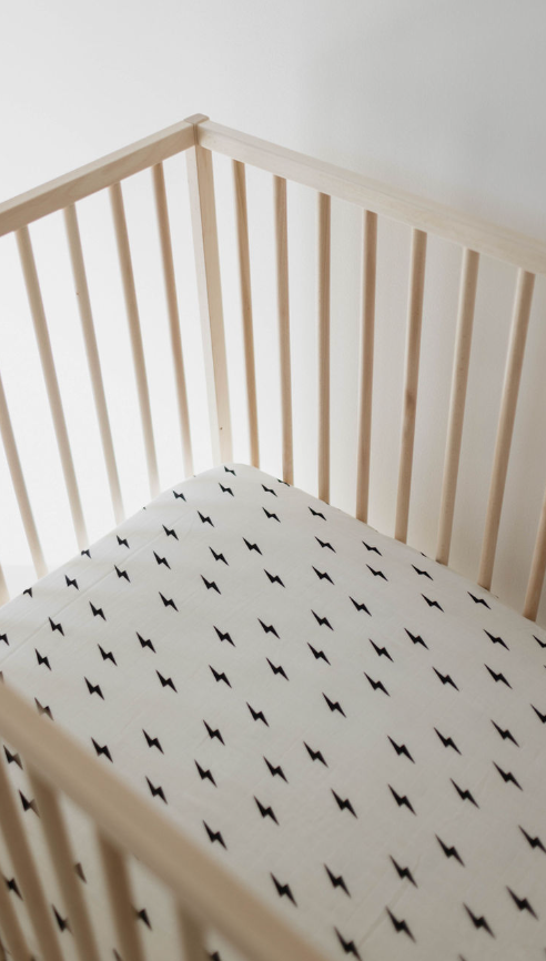 A minimalist wooden crib with slatted sides is showcased, featuring the Muslin Crib Sheet in White & Black Lightning Bolt from the forever french baby collection. Positioned against a white wall, it exudes a clean and modern aesthetic.