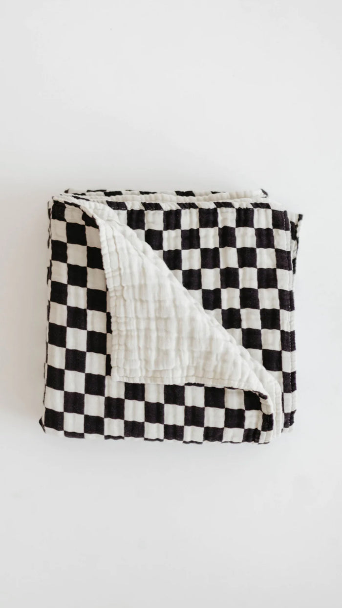 A folded Muslin Quilt | Black & White Checkerboard from forever french baby with one corner flipped to reveal a plain white backside sits on a white background. Made from soft muslin cotton, the quilt is neatly arranged, showcasing its contrasting patterns and textures, perfect for adding charm to your Forever French Baby collection.