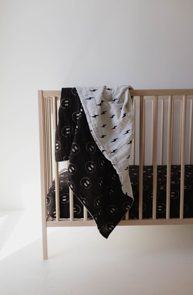 A minimalist wooden crib stands against a white wall. Draped over the crib's side are two blankets from Forever French Baby: one black with white circular patterns and another, the Muslin Quilt | ff Smile Black & White Bolt, with a gray background and black lightning bolt designs. The crib mattress also has a matching black cover with white patterns.