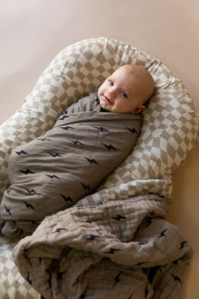 A baby swaddled in the Muslin Quilt | Lightning Bolt & Wavy Checker from forever french baby, featuring a 47x47 inch muslin cotton design with small black lightning bolt patterns, rests on a checkered cushion. The baby is looking up with a calm expression. The neutral-colored background complements the tones of the Forever French Baby blanket and cushion perfectly.