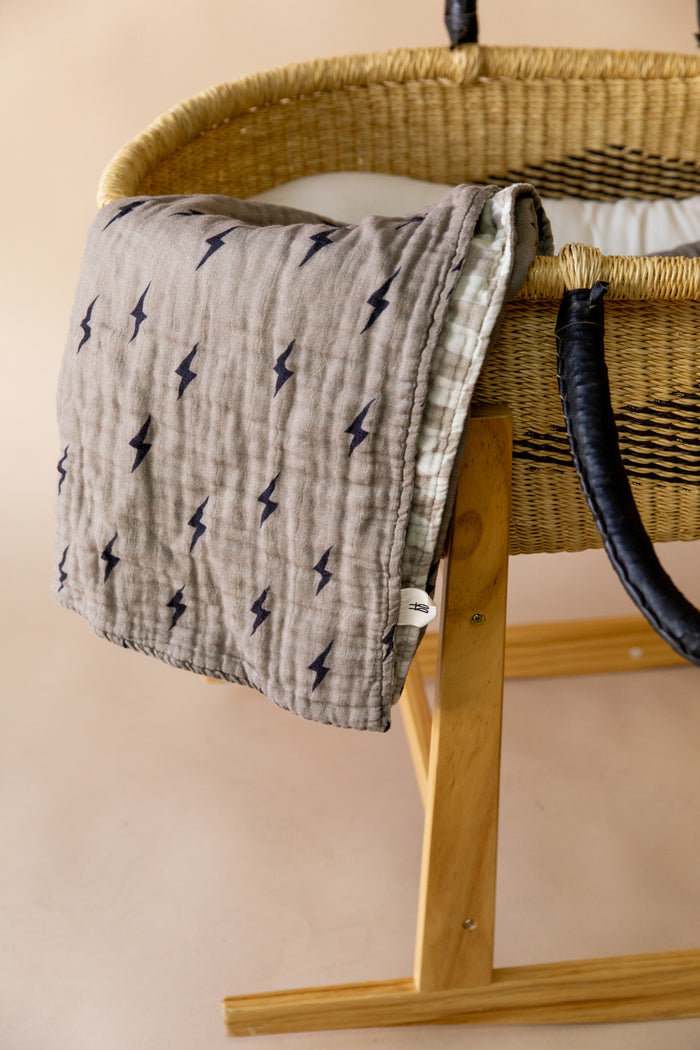 A woven baby bassinet with black handles stands on a wooden stand. A Muslin Quilt | Lightning Bolt & Wavy Checker by forever french baby, sized at 47x47 inches, is draped over the side of the bassinet. The background is a soft beige color, complementing the elegant style of Forever French Baby.